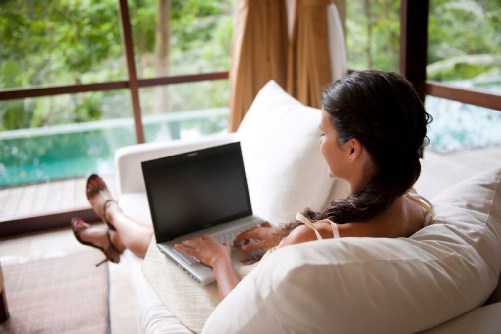 Woman relaxing on a sofa while typing on a laptop near a pool, showing the flexibility and freedom of freelance writing remote jobs for beginners.