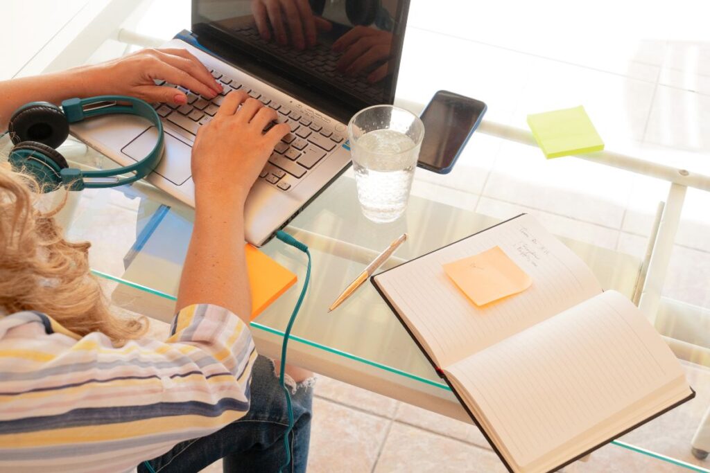 Close-up of a person typing on a laptop with headphones, notebook, and glass of water on a glass desk, showing a setup for online chat support remote jobs.