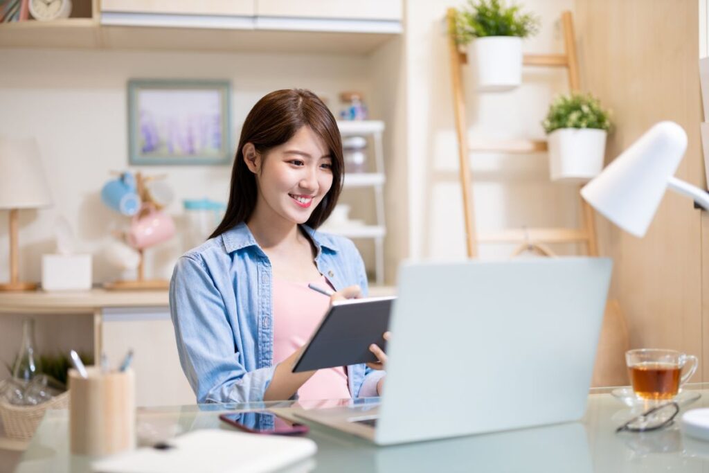 Smiling woman taking notes while working on a laptop at home, showing a productive setup for remote jobs for beginners and work-from-home jobs with no experience.