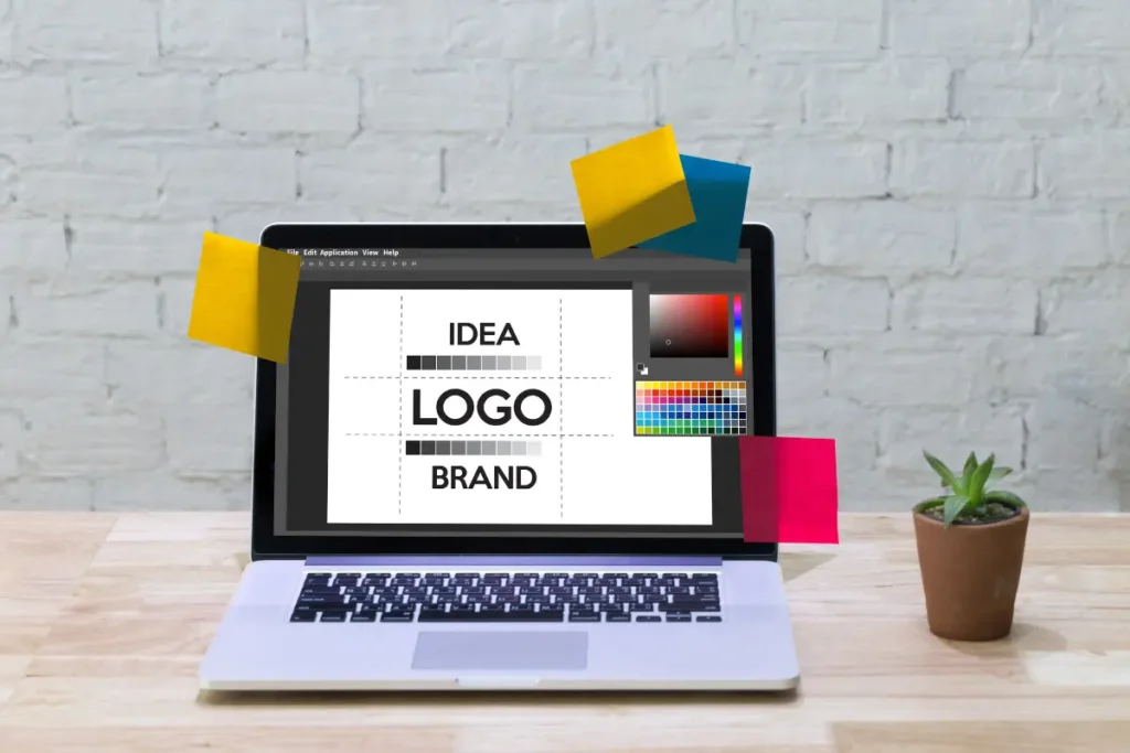 A laptop on a wooden desk displaying a graphic design program with the words “IDEA,” “LOGO,” and “BRAND” on the screen, surrounded by colorful sticky notes and a small potted plant, representing creative work-from-home jobs for beginners in design and branding.