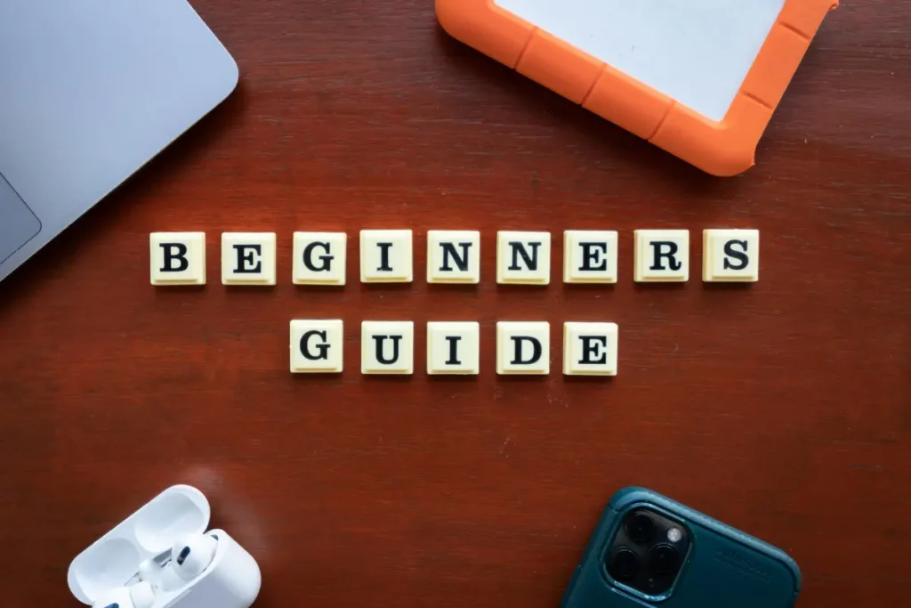 Scrabble-style tiles spelling “BEGINNERS GUIDE” on a wooden desk surrounded by a laptop, smartphone, tablet, and wireless earbuds, symbolizing a starter guide.